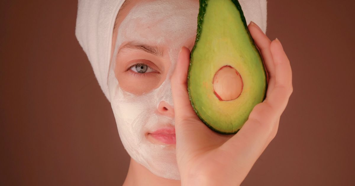 Face of woman with face mask holding avodcado half in her hand in front of one eye