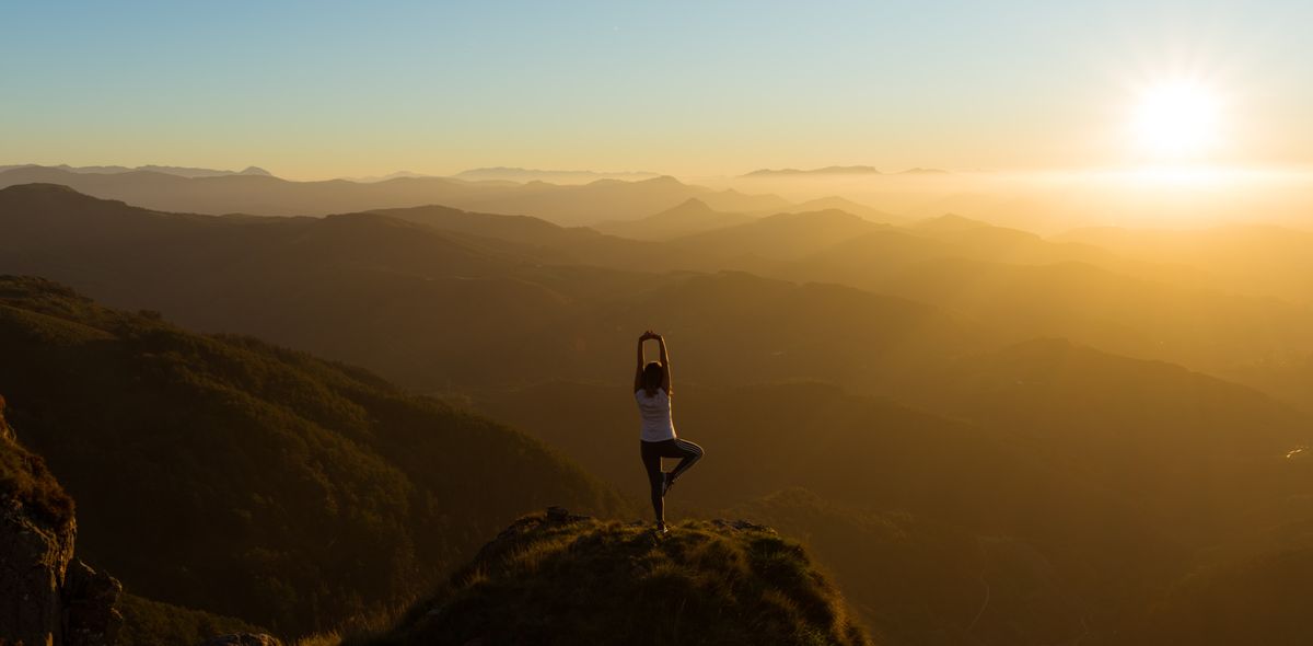 Woman on top of mountain practising the yoga asana the tree with view over the summits during sunset